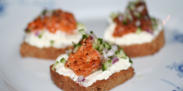 Ristet brød med rygeostcreme og varmrøget laks samt agurk og rødløg på toppen.